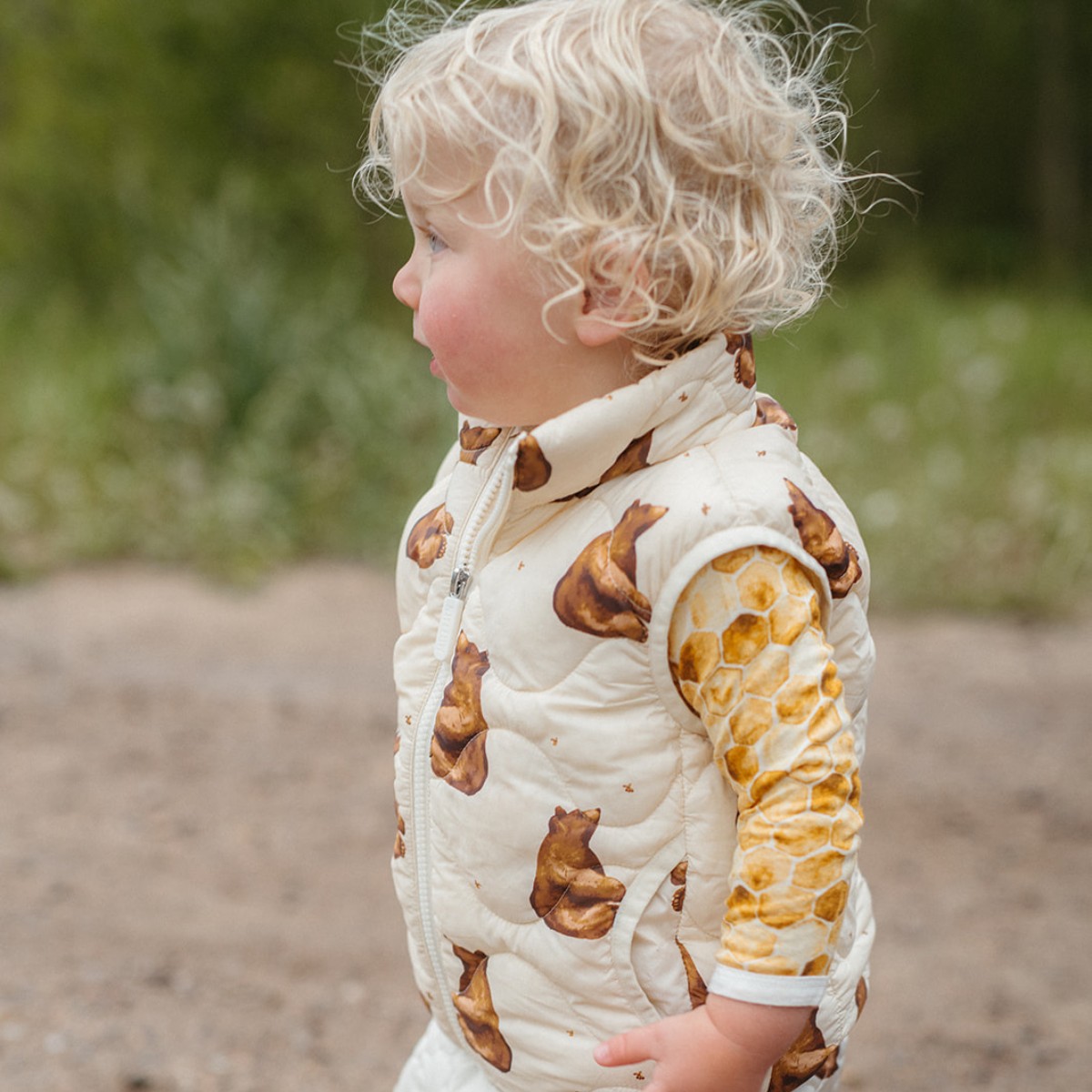 Profile of a baby or toddle outside wearing the honey bear lightweight quilted down vest with a bamboo long sleeve one pice underneath in the honeycomb print