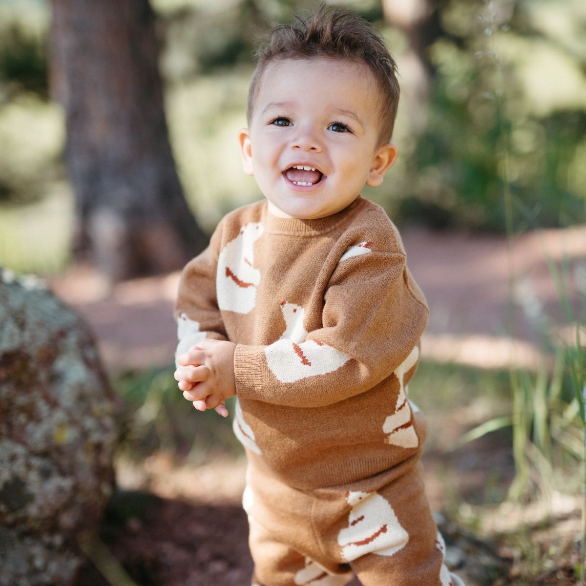 Baby or toddler in the forest holding his hands while smiling and wearing the Knitted Birdseye Jacquard Sweater and sweatpants