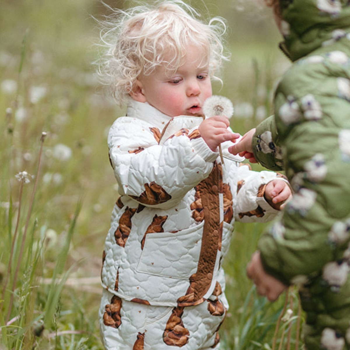 Baby and toddler in a green field of dandelions blowing on a dandelion while wearing the honey bear Milkbarn Quilted Jacket and Scarf